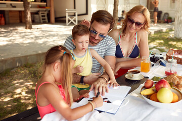 daughter showing to a little brother how to draw at outdoor table, having breakfast