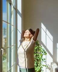 Enjoying new day. Beautiful happy mature woman stretching body and smiling while standing with closed eyes in sun rays shining through window on sunny morning at home. Optimism and happiness concept