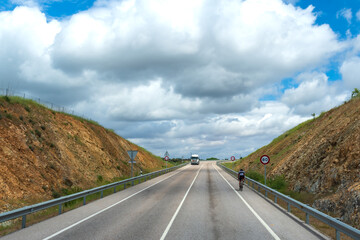 Fototapeta premium Road with a bicyclist on the shoulder and a truck in the opposite lane driving, with a cloudy sky.
