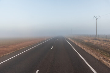 Straight road on a plain with fog in the background and poles with electrical wiring on the edge.