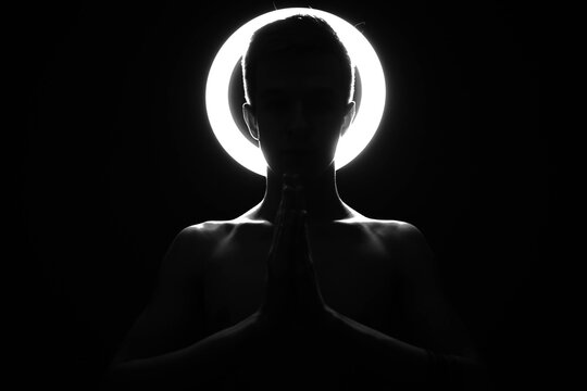 A silhouette of a man against the background of a shining halo, who, with folded hands, prays. Beautiful clavicle in black and white in