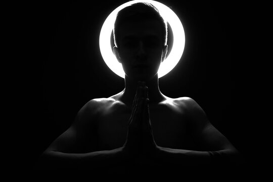 A silhouette of a man against the background of a shining halo, who, with folded hands, prays. Beautiful clavicle in black and white in