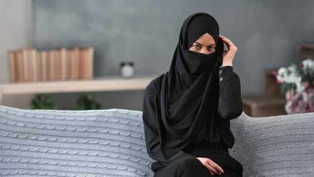 Mystery Arabic woman hiding face under niqab relaxing on couch at comfortable living room 