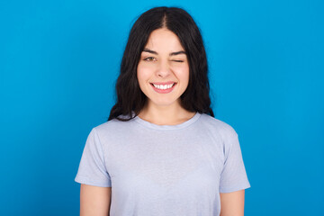 Obraz premium Coquettish young beautiful tattooed girl wearing blue t-shirt standing against blue background smiling happily, blinking at camera in a playful manner, flirting with you.