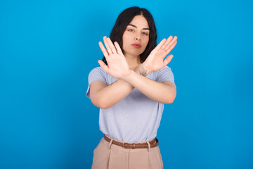 young beautiful tattooed girl wearing blue t-shirt standing against blue background has rejection expression crossing arms and palms doing negative sign, angry face.