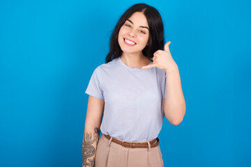 Fototapeta premium young beautiful tattooed girl wearing blue t-shirt standing against blue background smiling doing phone gesture with hand and fingers like talking on the telephone. Communicating concepts.