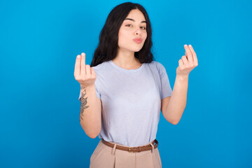 Fototapeta premium young beautiful tattooed girl wearing blue t-shirt standing against blue background doing money gesture with hands, asking for salary payment, millionaire business