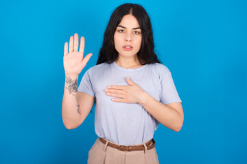 Fototapeta premium young beautiful tattooed girl wearing blue t-shirt standing against blue background Swearing with hand on chest and open palm, making a loyalty promise oath