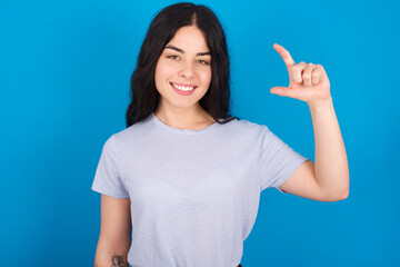 young beautiful tattooed girl wearing blue t-shirt standing against blue background smiling and confident gesturing with hand doing small size sign with fingers looking and the camera. Measure concept