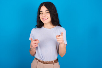 Fototapeta premium young beautiful tattooed girl wearing blue t-shirt standing against blue background pointing fingers to camera with happy and funny face. Good energy and vibes.