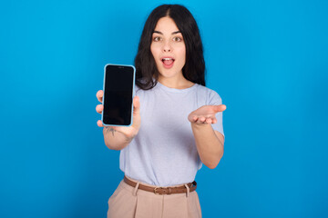 young beautiful tattooed girl wearing blue t-shirt standing against blue background with a mobile. presenting smartphone. Advertisement concept.