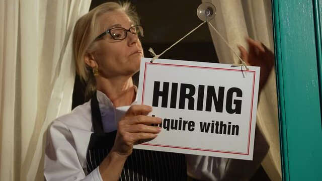 Female shop owner putting Help Wanted sign on glass door. Woman staff chef of restaurant advertising for hiring staff.