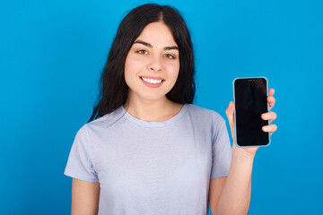 Fototapeta premium Smiling young beautiful tattooed girl wearing blue t-shirt standing against blue background Mock up copy space. Hold mobile phone with blank empty screen