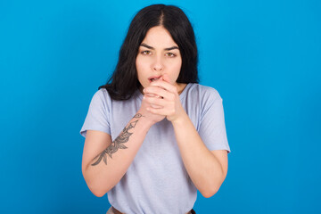 young beautiful tattooed girl wearing blue t-shirt standing against blue background holding oneself, feels very cold outside, hopes that will not get cold