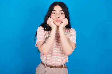 young beautiful tattooed girl wearing pink striped t-shirt standing against blue background with surprised expression keeps hands under chin keeps lips folded makes funny grimace