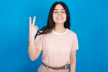 Fototapeta premium young beautiful tattooed girl wearing pink striped t-shirt standing against blue background smiling and looking friendly, showing number two or second with hand forward, counting down