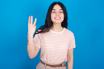 Fototapeta premium young beautiful tattooed girl wearing pink striped t-shirt standing against blue background smiling and looking friendly, showing number three or third with hand forward, counting down