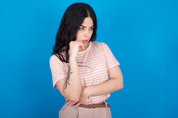 Astonished young beautiful tattooed girl wearing pink striped t-shirt standing against blue background looks aside surprisingly with opened mouth.