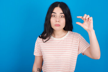 Fototapeta premium young beautiful tattooed girl wearing pink striped t-shirt standing against blue background purses lip and gestures with hand, shows something very little.