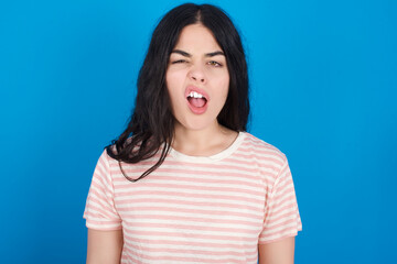 young beautiful tattooed girl wearing pink striped t-shirt standing against blue background yawns with opened mouth stands. Daily morning routine