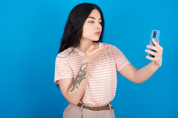 young beautiful tattooed girl wearing pink striped t-shirt standing against blue background blows air kiss at camera of smartphone and takes selfie, sends mwah via online call.
