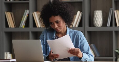 Young African businesswoman sit at desk check documents working on laptop, holding sheets reading written text agreement, writing on device. Paperwork, pay bill through e-banking application concept