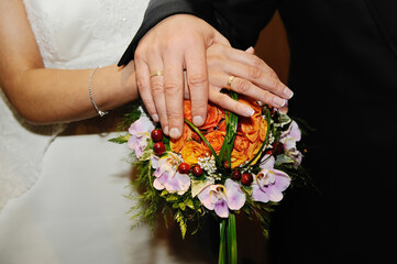 Gold rings on the joined hands of the newlyweds on the bride's bouquet of orange flowers.