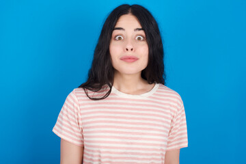 Obraz premium Stunned young beautiful tattooed girl wearing pink striped t-shirt standing against blue background stares reacts on shocking news. 