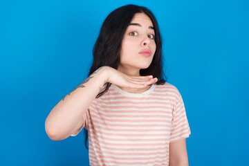 young beautiful tattooed girl wearing pink striped t-shirt standing against blue background cutting throat with hand as knife, threaten aggression with furious violence.