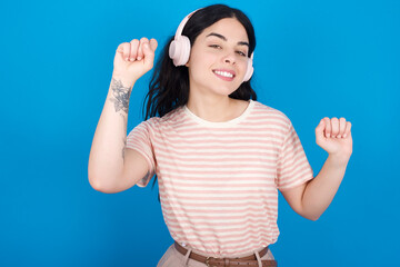 Fototapeta premium Carefree young beautiful tattooed girl wearing pink striped t-shirt thick beard and toothy smile raises arms dances carefree moves with rhythm of music listens music from playlist via headphones