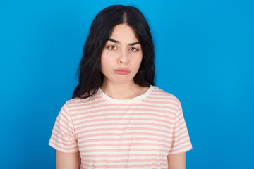Obraz premium Displeased young beautiful tattooed girl wearing pink striped t-shirt standing against blue background frowns face feels unhappy has some problems. Negative emotions and feelings concept