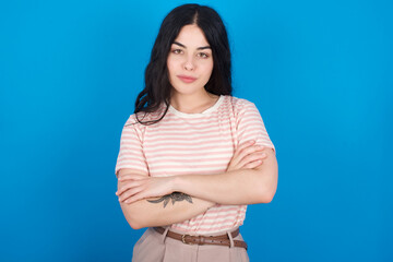 Confident young beautiful tattooed girl wearing pink striped t-shirt standing against blue background with arms crossed looking to the camera
