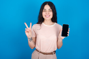 young beautiful tattooed girl wearing pink striped t-shirt standing against blue background holding modern device showing v-sign