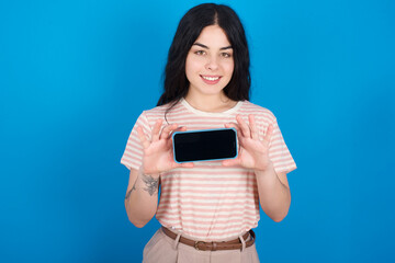 Cheerful cheery content young beautiful tattooed girl wearing pink striped t-shirt standing against blue background holding in hands smartphone with blank space screen 
