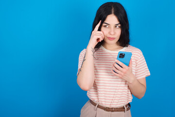 young beautiful tattooed girl wearing pink striped t-shirt standing against blue background holding gadget while sticking out tongue