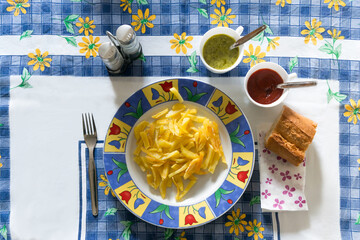 Plate of freshly made French fries set out on a colorful table alongside two sauces, salt, pepper, and bread.