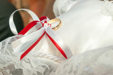 Gold ring knotted with a red and white bow placed on a white fabric cushion.