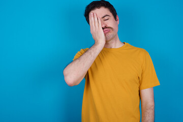 Tired overworked young handsome Caucasian man with moustache wearing orange t-shirt  has sleepy expression, gloomy look, covers face with hand, has eyes shut, gasps from tiredness, fatigue after party