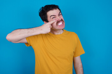 young handsome Caucasian man with moustache wearing orange t-shirt against blue background pointing unhappy to pimple on forehead, ugly infection of blackhead. Acne and skin problem