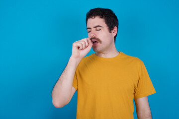 young handsome Caucasian man with moustache wearing yellow t-shirt against blue background feeling unwell and coughing as symptom for cold or bronchitis. Healthcare concept.