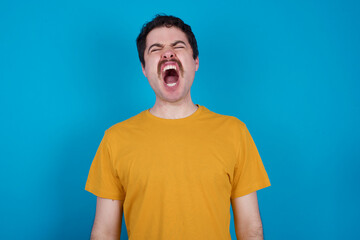 young handsome Caucasian man with moustache wearing yellow t-shirt against blue background angry...