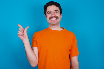 young handsome Caucasian man with moustache wearing orange t-shirt against blue background pointing up with fingers number eight in Chinese sign language BÄ.