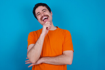 young handsome Caucasian man with moustache wearing orange t-shirt against blue background laughs...