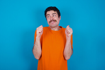 young handsome Caucasian man with moustache wearing orange t-shirt against blue background clenches fists and awaits for something nice happened looks away bites lips and waits announcement of results