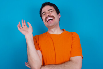 Overjoyed successful young handsome Caucasian man with moustache wearing orange t-shirt against blue background raises palm and closes eyes in joy being entertained by friends