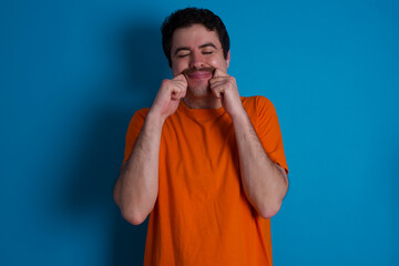 Pleased young handsome Caucasian man with moustache wearing orange t-shirt against blue background...