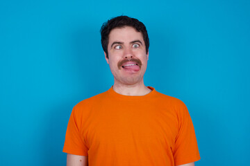 Fototapeta premium Funny young handsome Caucasian man with moustache wearing orange t-shirt against blue background makes grimace and crosses eyes plays fool has fun alone sticks out tongue.