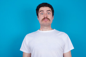 Stunned young handsome Caucasian man with moustache wearing white t-shirt against blue background stares reacts on shocking news. Astonished male holds breath