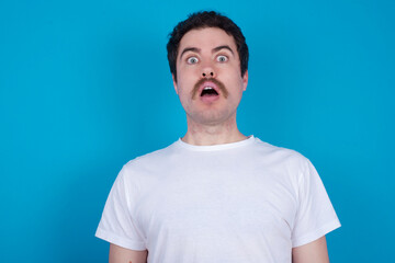 Fototapeta premium Shocked young handsome Caucasian man with moustache wearing white t-shirt against blue background stares bugged eyes keeps mouth opened has surprised expression. Omg concept