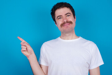 Fototapeta premium young handsome Caucasian man with moustache wearing white t-shirt against blue background points to side on blank space demonstrates advertisement. People and promotion concept